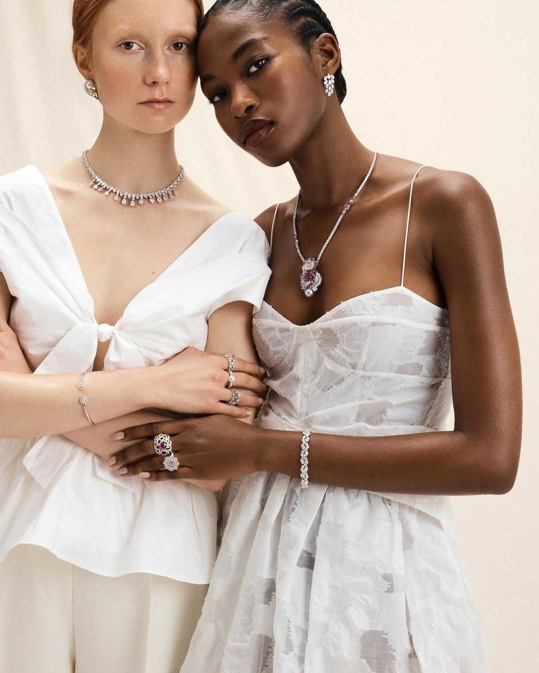 Two fashion models pose indoors wearing white tops and dresses, adorned with diamond jewelry, including necklaces, earrings, rings, and bracelets.