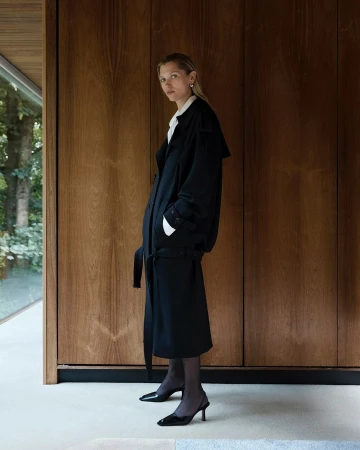 Woman in a black belted trench coat standing against dark wood paneling in an interior space