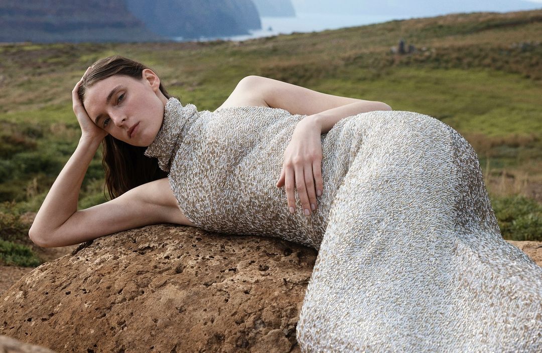A woman in an embellished gown poses lying down on a rock formation with a soft focus of cliffs and water in the background.