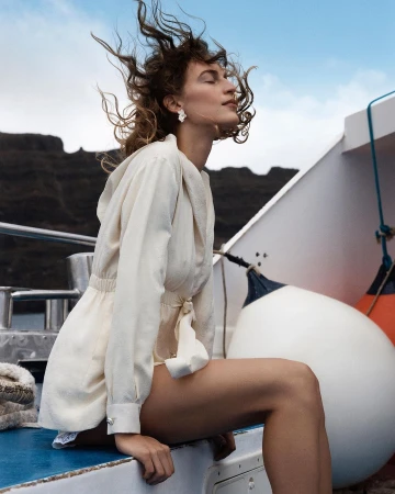 Woman with wind-blown brown hair seated on a boat wearing a cream-coloured belted jacket against an ocean and rocky coast background