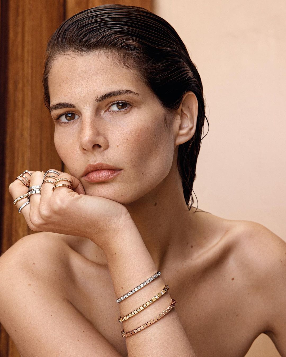 Close-up studio portrait of a beautiful woman with wet styled hair, wearing bracelets and rings, set against a tan backdrop.