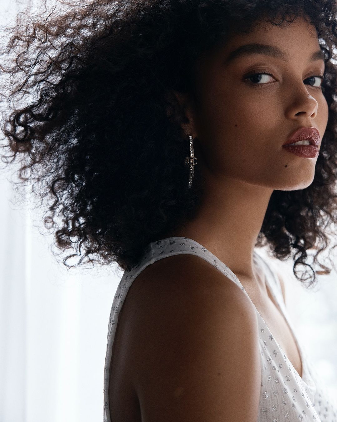 Portrait of a young Black woman with curly hair wearing a white sleeveless top and statement earrings against a bright white background.
