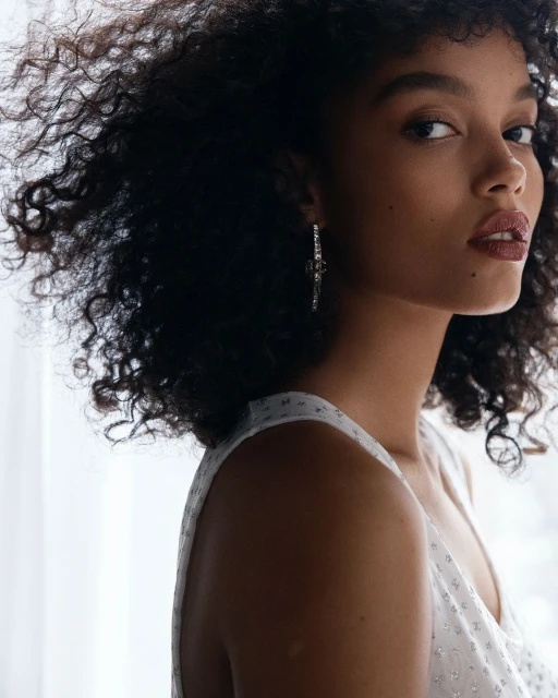 Portrait of a young Black woman with curly hair wearing a white sleeveless top and statement earrings against a bright white background.