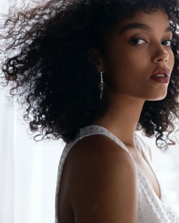 Portrait of a young Black woman with curly hair wearing a white sleeveless top and statement earrings against a bright white background.