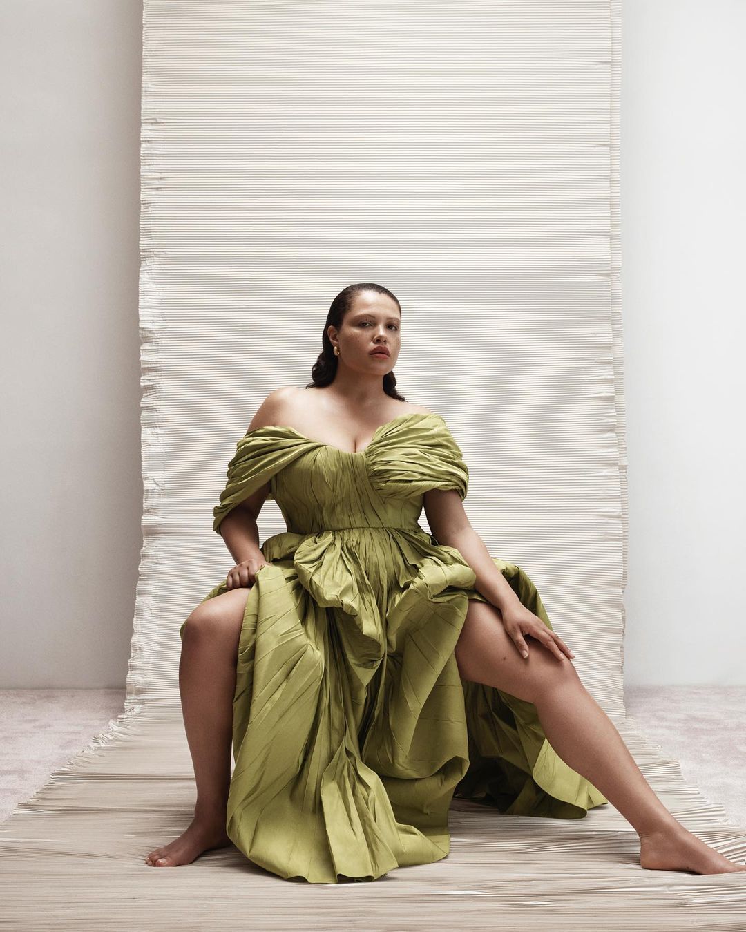 Woman in green gown seated in photo studio on textured floor covering. Studio shot showing pleated fabric backdrop and soft lighting.