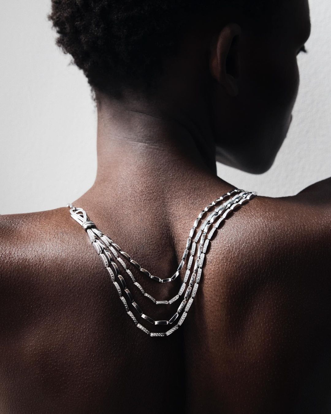 A close-up of a woman's bare back and shoulders wearing a silver necklace. The woman has dark skin and is facing away from the camera.