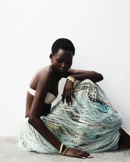 A black woman poses in a fringed skirt and bra top against a white wall. She is adorned with gold bracelets and earrings.