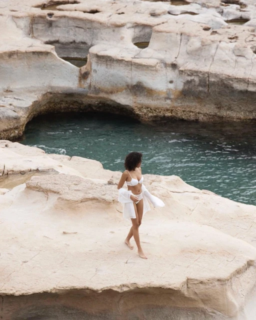 Woman in bikini and white cover-up standing on rocky shore near ocean. Blue water and rock formations create a serene backdrop.