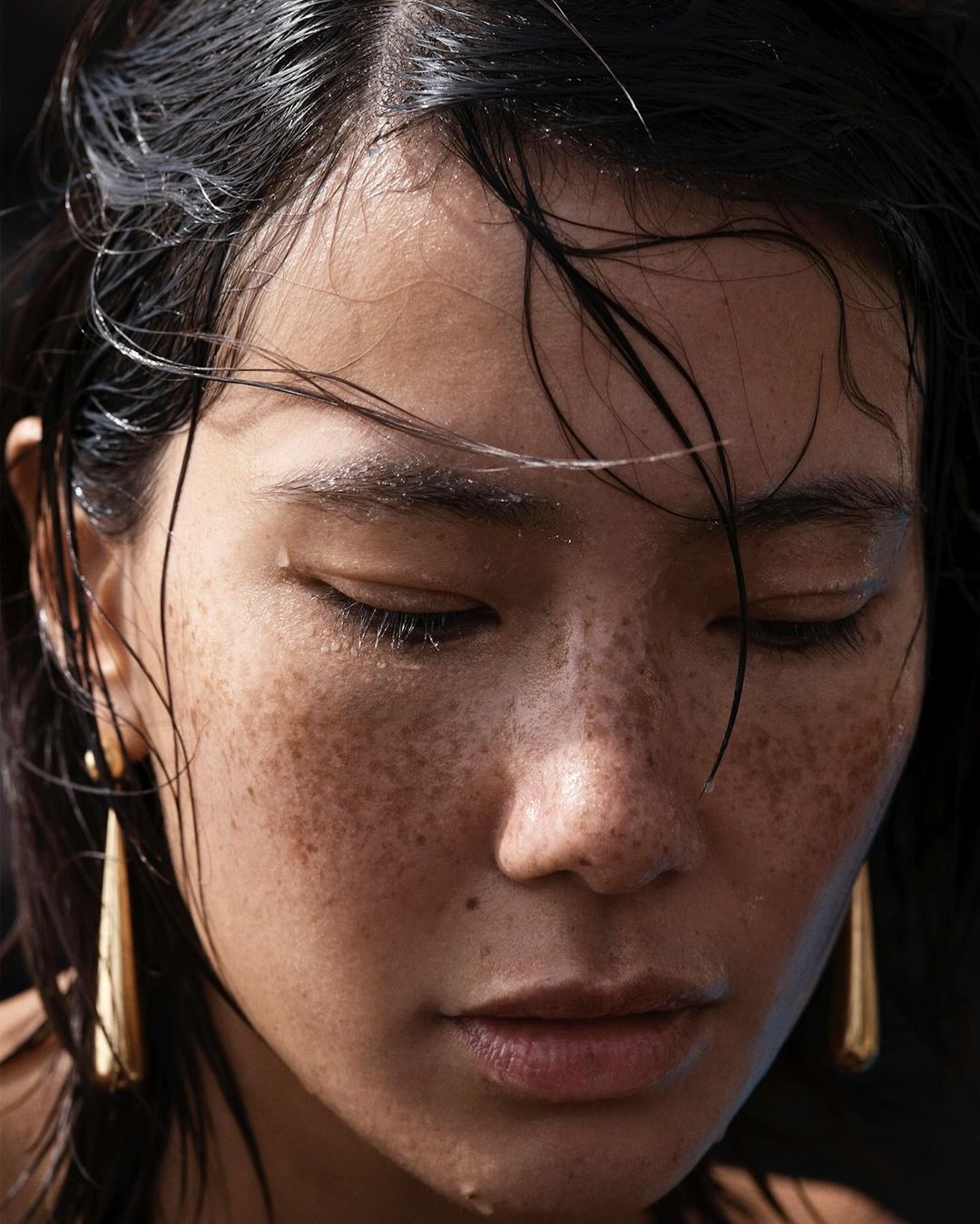 Close-up portrait of a woman with dark hair and dewy skin, adorned with gold teardrop earrings, capturing a serene and intimate moment.