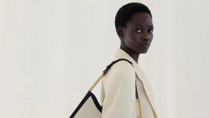 Black woman with short hair wearing a cream blazer and carrying a structured shoulder bag against a plain white background