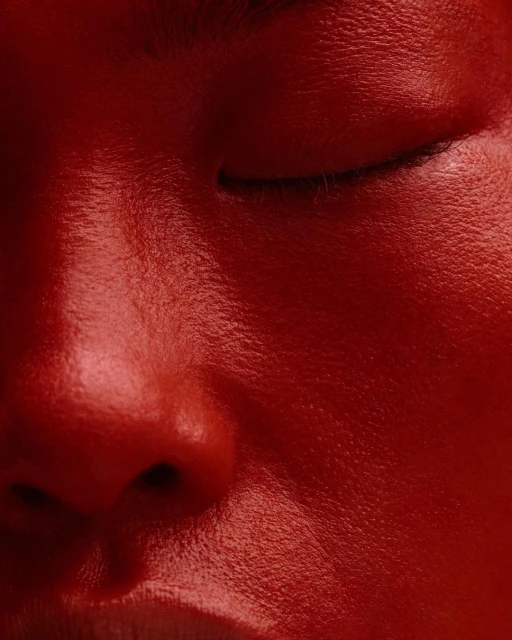 Close-up of an adult human's face, eyes closed, with red color grading, capturing skin texture and facial features in a studio setting.