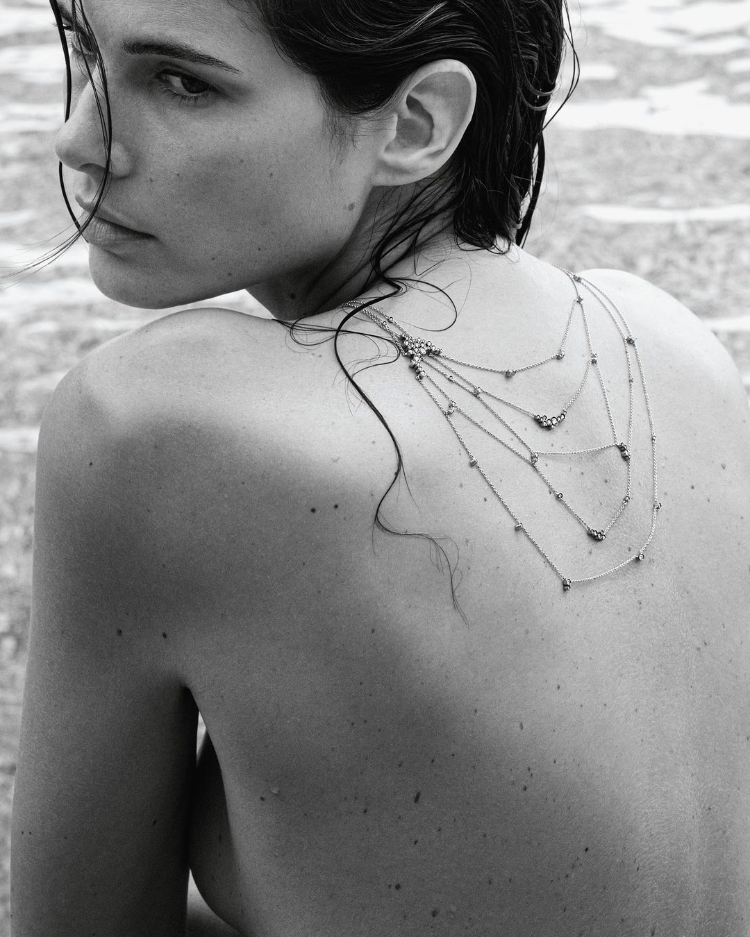 Monochrome portrait of a woman with layered necklaces, looking over her shoulder in a neutral outdoor setting with light reflecting on water.