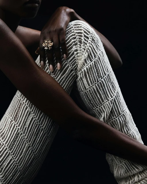 Close-up of an adult woman with dark skin wearing a gold ring and white patterned pants in a studio setting with a dark background.