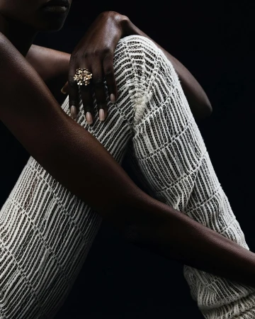 Close-up of an adult woman with dark skin wearing a gold ring and white patterned pants in a studio setting with a dark background.