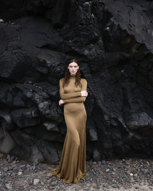 A woman in a long olive dress poses against a textured black rock formation outdoors on a beach with small gray rocks in soft lighting.