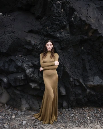 A woman in a long olive dress poses against a textured black rock formation outdoors on a beach with small gray rocks in soft lighting.