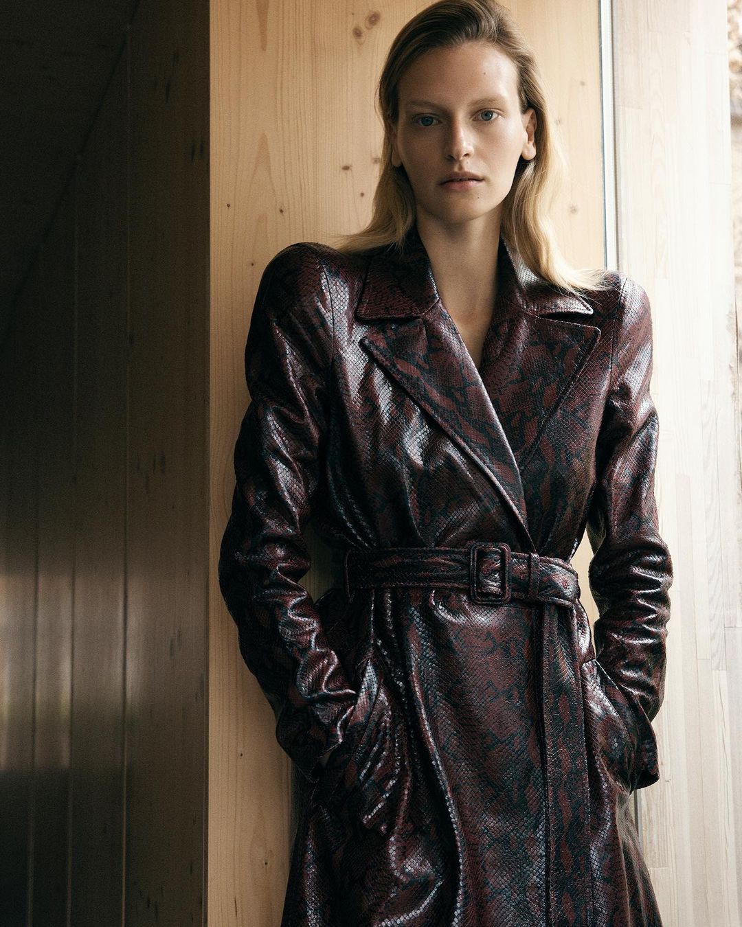 A blonde woman poses in a textured snakeskin trench coat indoors, with a serious expression, under natural light.