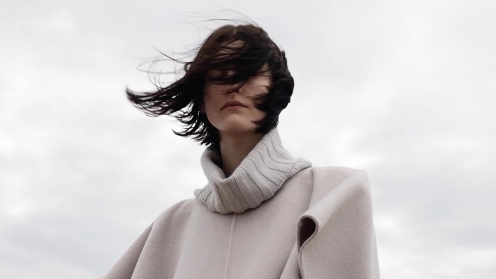 A woman with short brown hair wearing a turtleneck and light coat stands against a cloudy sky, her hair blowing in the wind.
