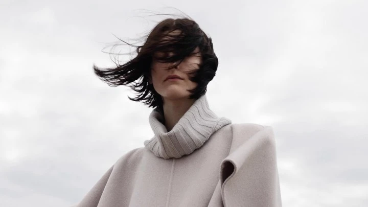 A woman with short brown hair wearing a turtleneck and light coat stands against a cloudy sky, her hair blowing in the wind.