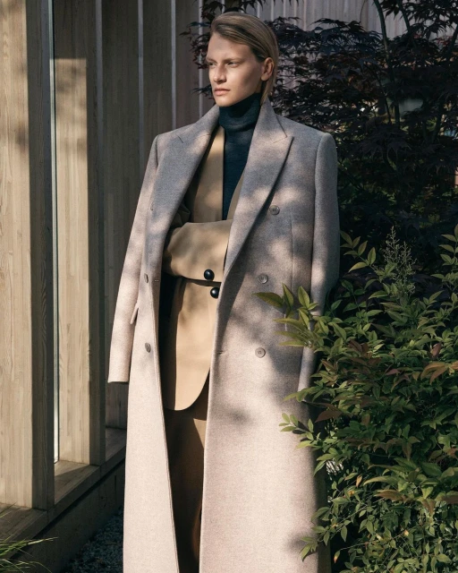Fashionable adult stands outdoors in tailored coat and suit with neutral tones, complemented by natural light and green foliage.