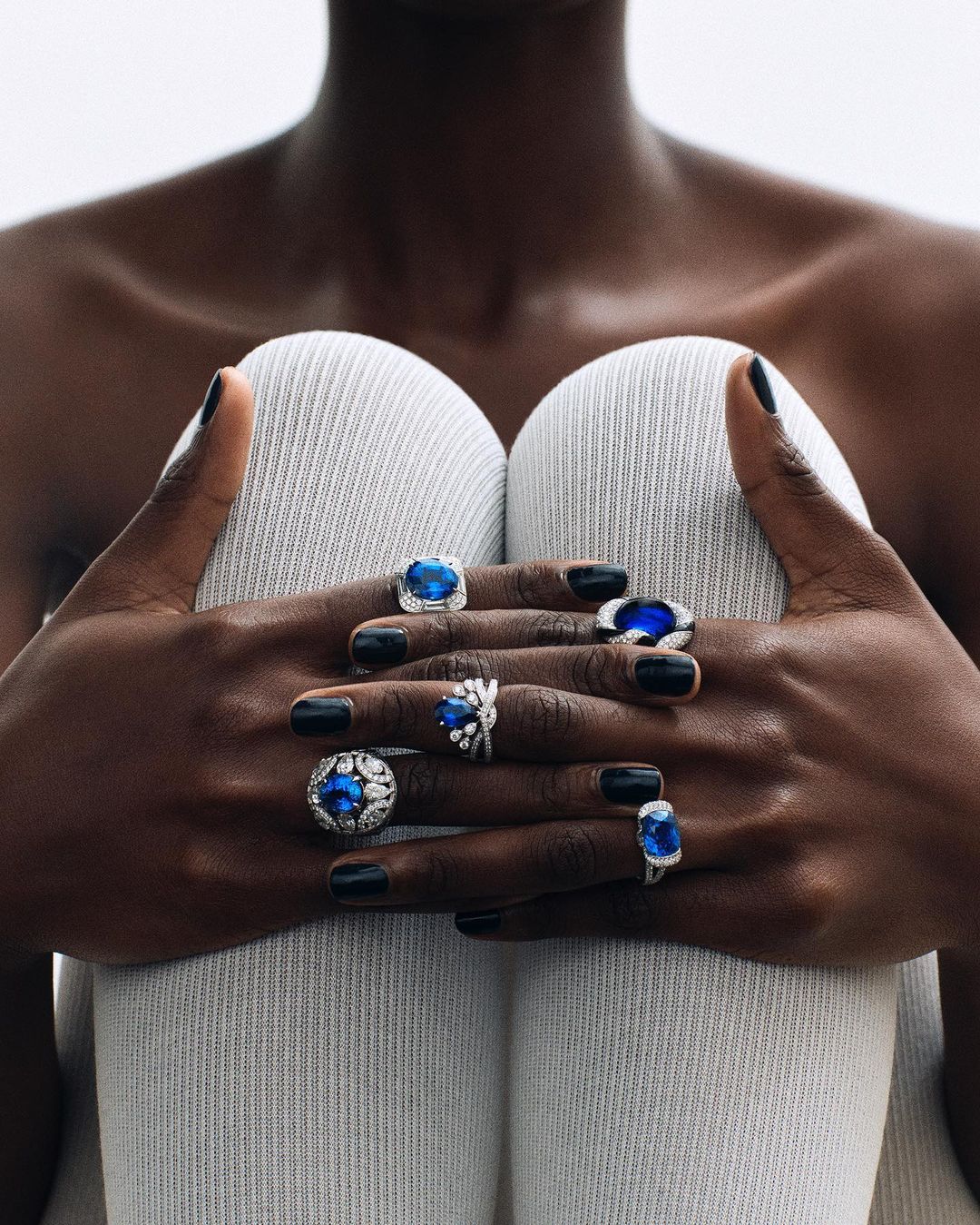 Close-up of dark-skinned adult with black nails wearing a variety of statement blue gemstone rings, holding white textured fabric.