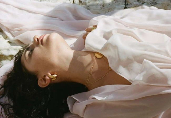 Model lying on light fabric wearing gold chain necklaces and earrings outdoors in sunlight