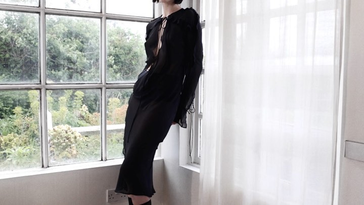 A slender woman in a black dress strikes a pose next to a bright window with sheer white curtains and a view of a green outdoor space.