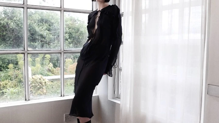 A slender woman in a black dress strikes a pose next to a bright window with sheer white curtains and a view of a green outdoor space.