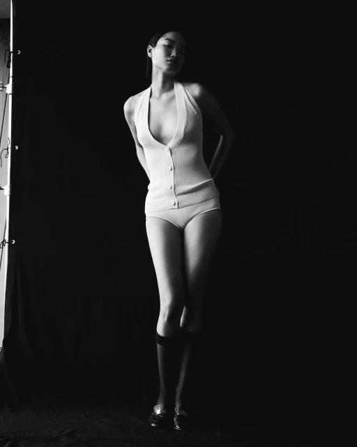 Monochrome fashion shot of a slender woman in a halter-top button vest, briefs, socks, and shoes against a dark background.