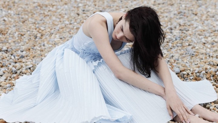 Brunette woman in pale blue dress sitting on pebble beach, head down, arms around legs, medium shot, soft lighting.