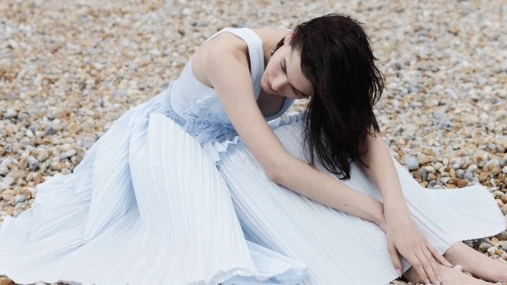 Brunette woman in pale blue dress sitting on pebble beach, head down, arms around legs, medium shot, soft lighting.