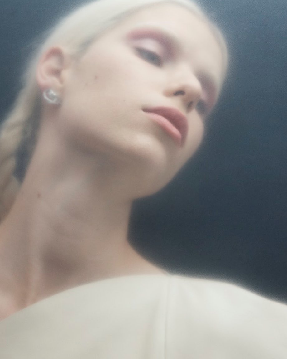Close-up portrait of a fair-skinned adult woman with blonde hair in braids, wearing an off-white top against a dark background.