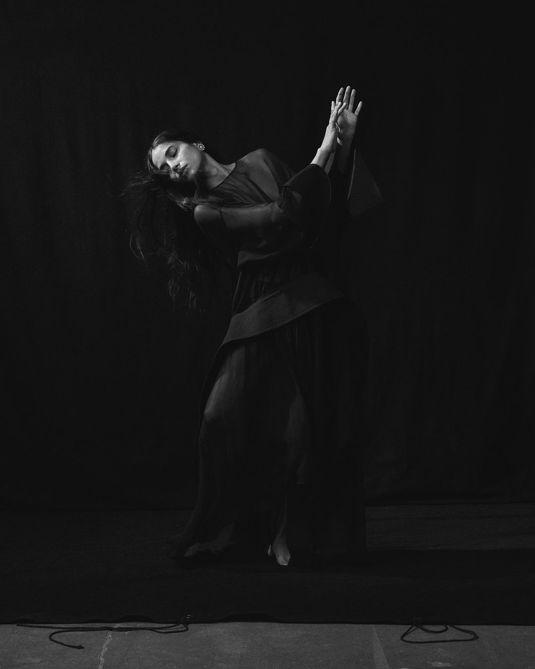 Monochrome portrait of a graceful woman in a flowing garment with outstretched hands, captured in a dance-like pose against a dark backdrop.