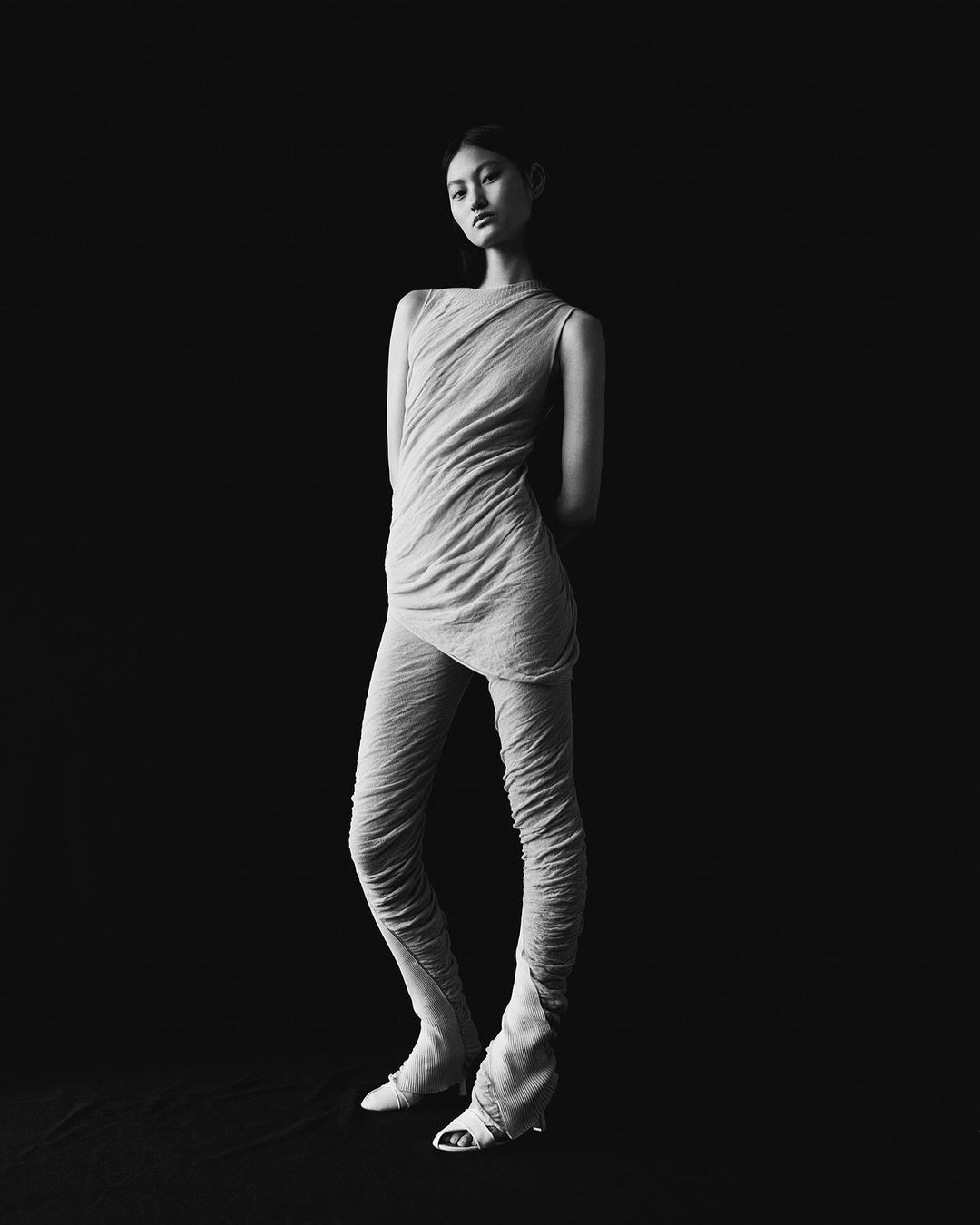 Monochrome fashion portrait of an Asian woman in a draped top and leggings with light-colored heels, set against a solid black background.