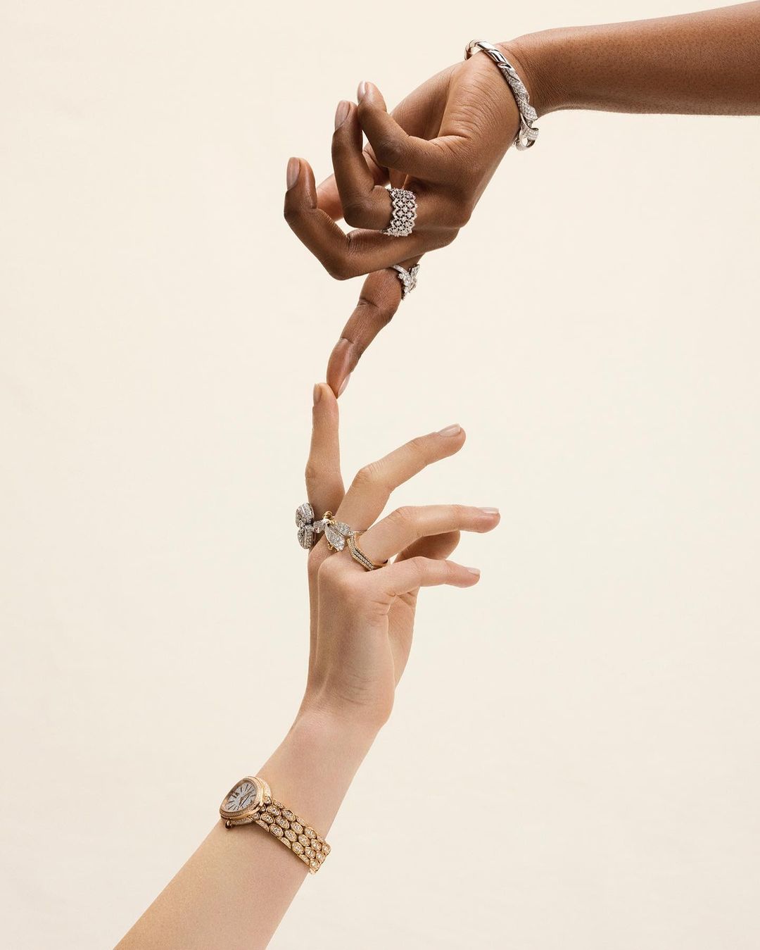 Two people's hands are touching, adorned with bracelets, rings, and a wristwatch, against a light beige backdrop. Close-up studio shot.