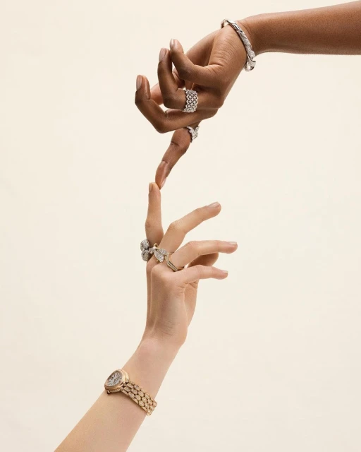 Two people's hands are touching, adorned with bracelets, rings, and a wristwatch, against a light beige backdrop. Close-up studio shot.