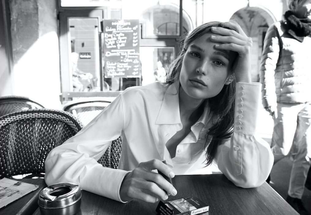 Black and white portrait of an adult female in a white blouse seated at a table with a cigarette, ashtray, and cigarettes in an indoor setting.