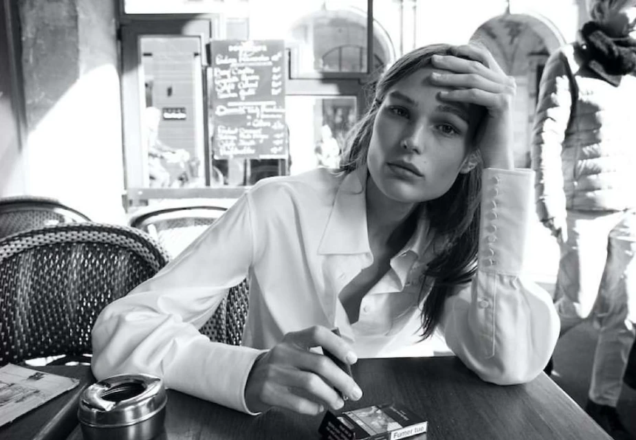 Black and white portrait of an adult female in a white blouse seated at a table with a cigarette, ashtray, and cigarettes in an indoor setting.