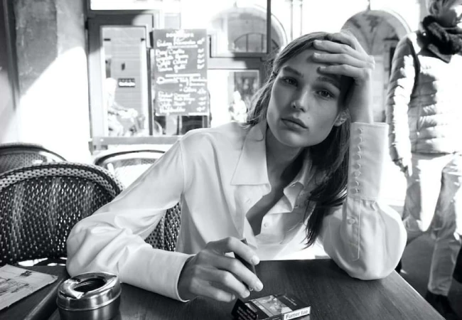 Black and white portrait of an adult female in a white blouse seated at a table with a cigarette, ashtray, and cigarettes in an indoor setting.