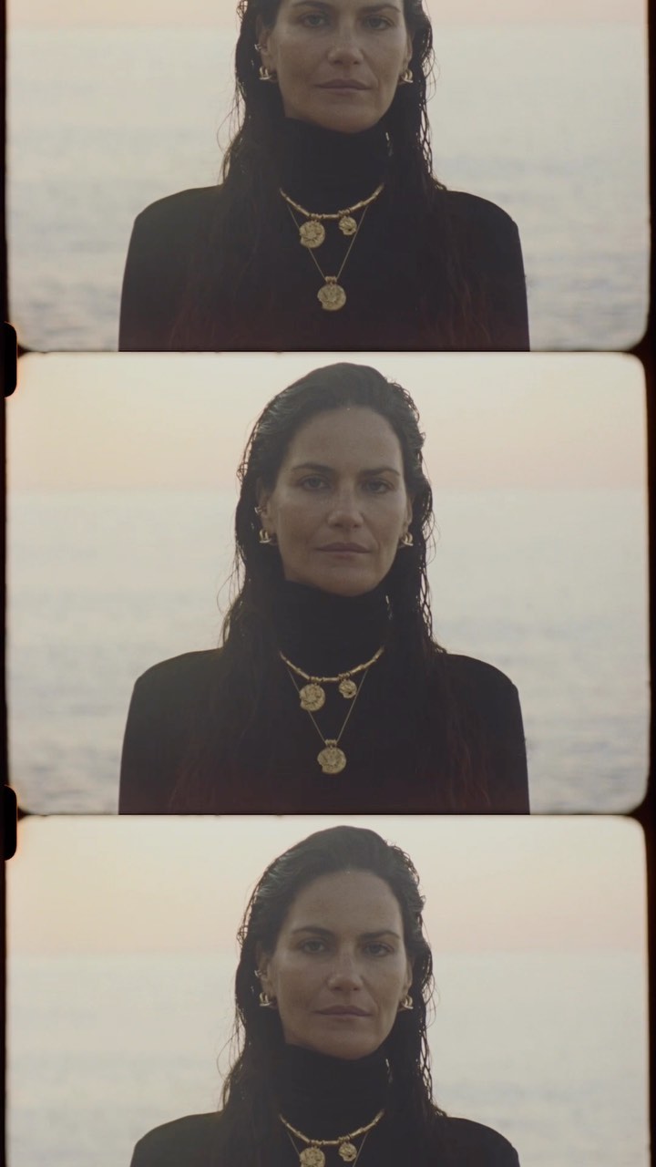 Close-up of an adult female model in front of an ocean backdrop, wearing a black top and gold jewelry, captured in a retro film style.
