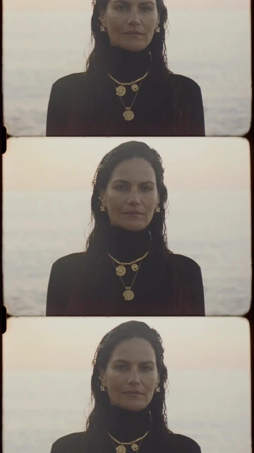 Close-up of an adult female model in front of an ocean backdrop, wearing a black top and gold jewelry, captured in a retro film style.