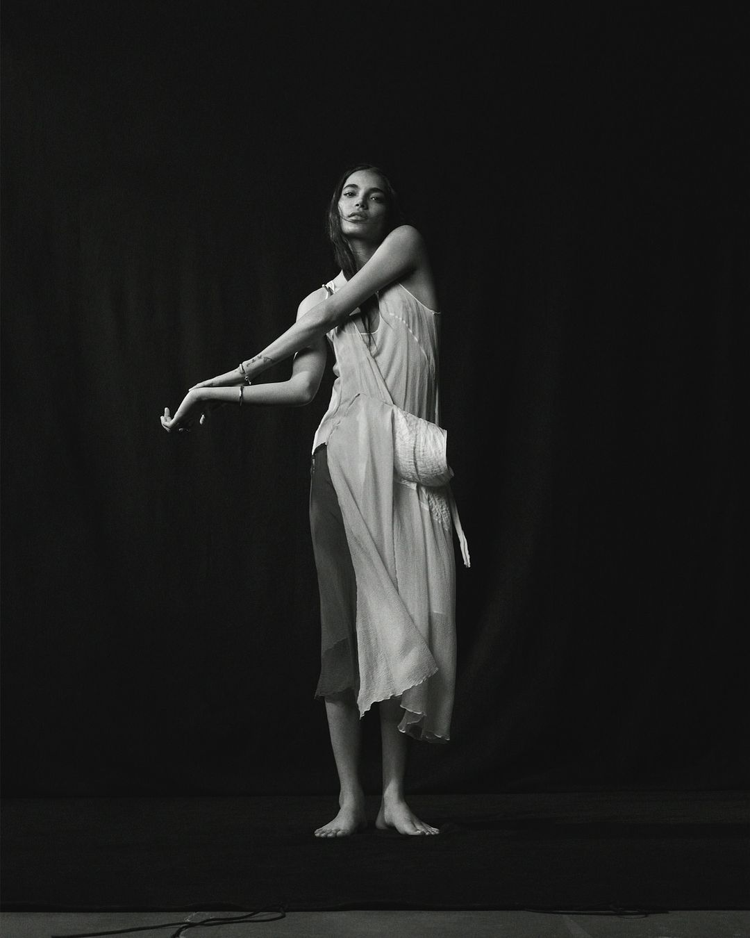 Monochrome fashion portrait of a young adult woman in a slip dress and shoulder bag in front of a dark background in a studio.