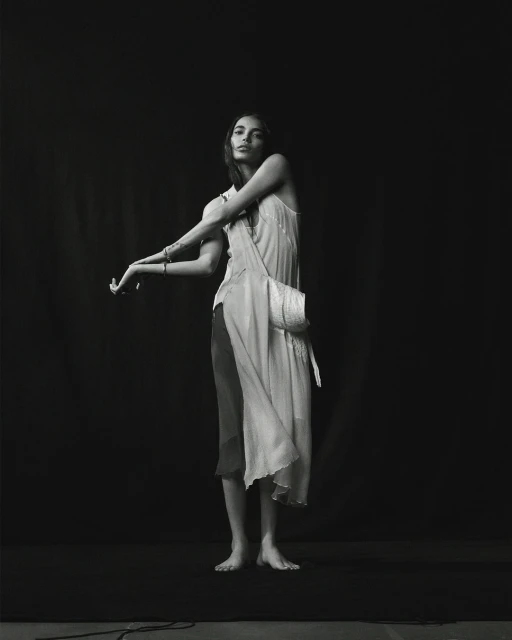 Monochrome fashion portrait of a young adult woman in a slip dress and shoulder bag in front of a dark background in a studio.