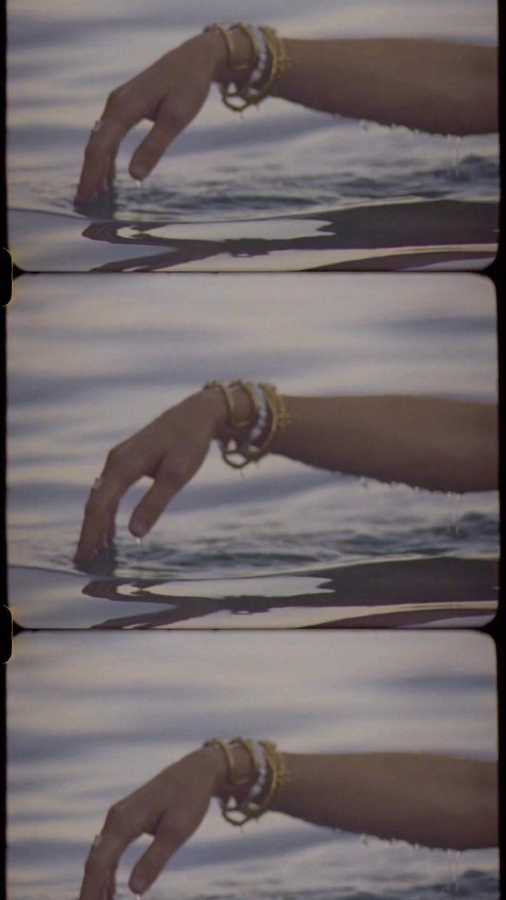 Close-up on an arm in water, adorned with bracelets. The water's surface reflects light in soft hues, creating a serene, ethereal atmosphere.