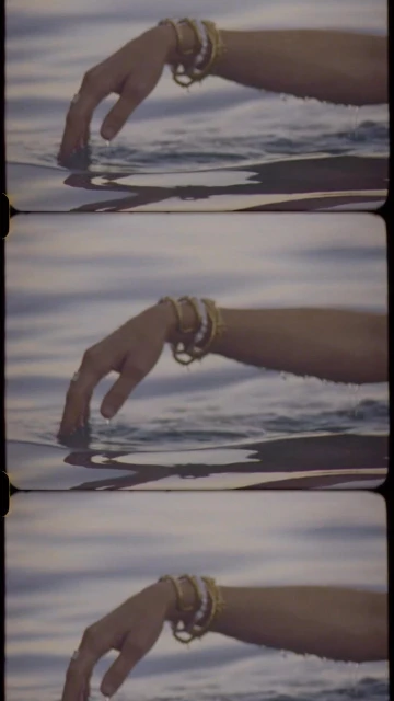 Close-up on an arm in water, adorned with bracelets. The water's surface reflects light in soft hues, creating a serene, ethereal atmosphere.