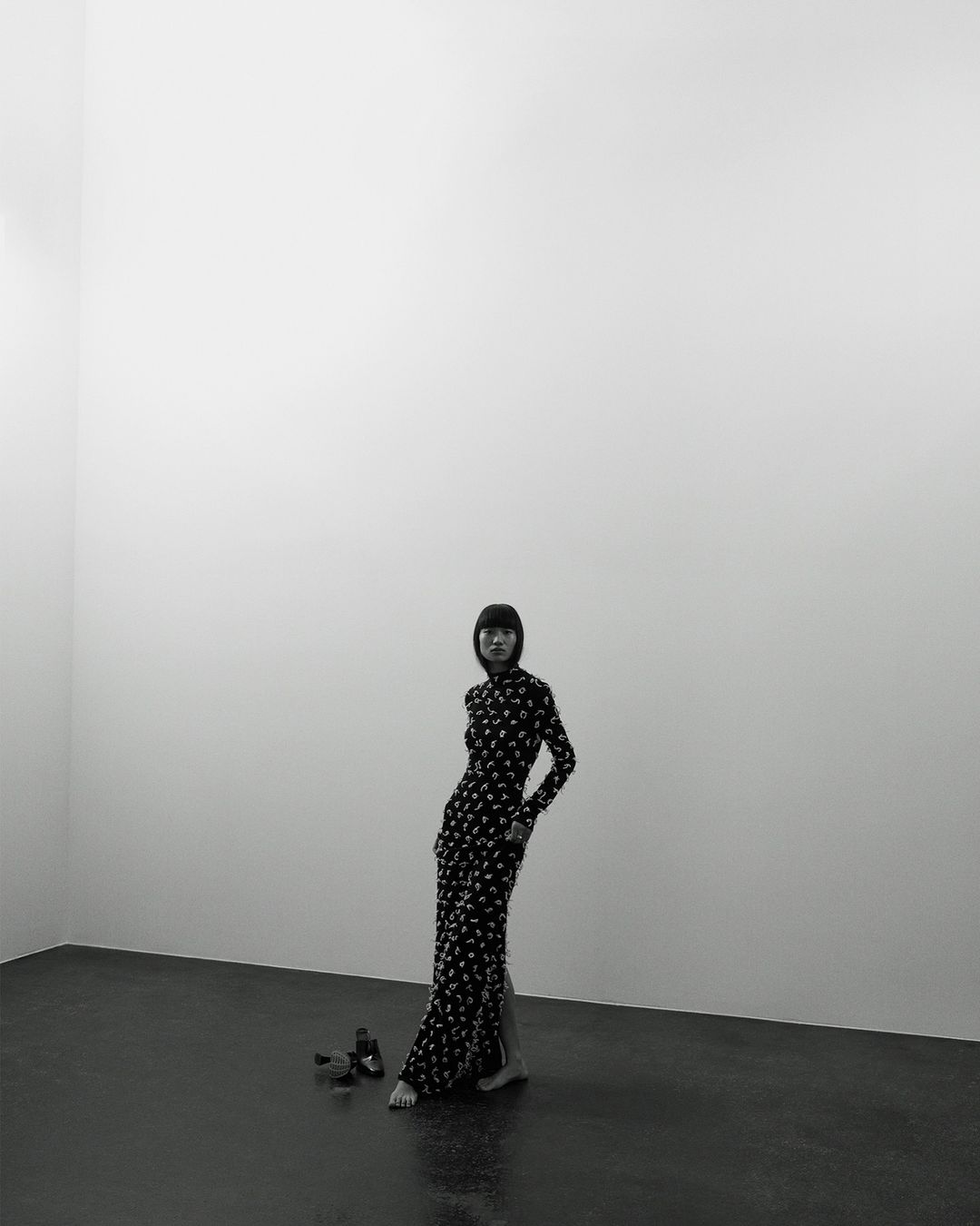 Fashion photo of a slender adult woman with dark hair wearing a long, patterned dress and posing barefoot in a stark, minimalist photo studio.
