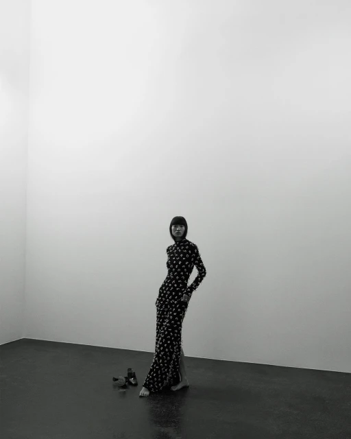 Fashion photo of a slender adult woman with dark hair wearing a long, patterned dress and posing barefoot in a stark, minimalist photo studio.
