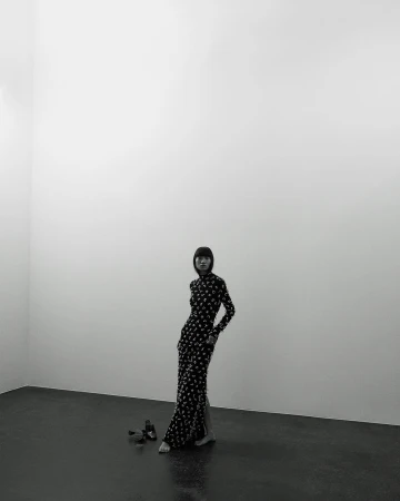 Fashion photo of a slender adult woman with dark hair wearing a long, patterned dress and posing barefoot in a stark, minimalist photo studio.