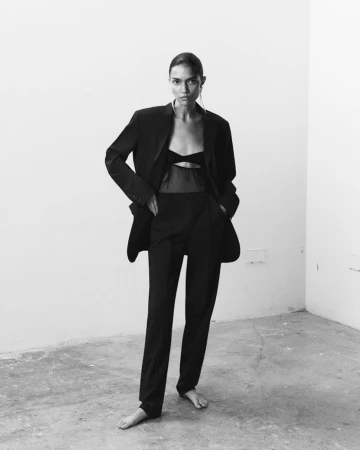 Model standing barefoot wearing an oversized black suit jacket and trousers against a neutral wall