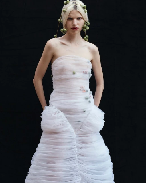 Fashion model in a strapless white gown with floral accents, wearing decorative headpiece against a dark backdrop in a studio setting.
